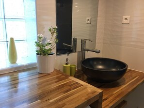 A modern bathroom with a wooden vanity, a black vessel sink, a white vase with flowers, and a mirror above the sink. - Quiet cottage + pool, for 3 pers. (Lézignan-Corbières)