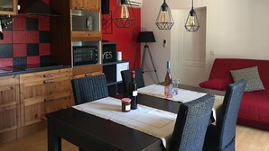 A modern kitchen with wooden cabinets, a built-in oven, and a red accent wall. There is a dining table with chairs and a red sofa in the living area. - Quiet cottage + pool, for 3 pers. (Lézignan-Corbières)