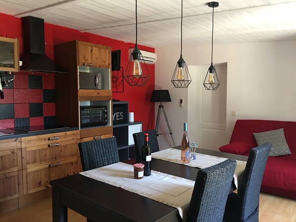 A modern kitchen with wooden cabinets, a built-in oven, and a red accent wall. There is a dining table with chairs and a red sofa in the living area. - Quiet cottage + pool, for 3 pers. (Lézignan-Corbières)