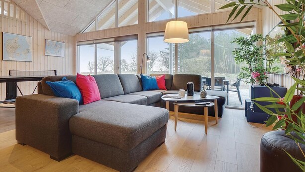 Un salon spacieux, doté d’un canapé d’angle, d’une table basse et d’un piano. De grandes fenêtres laissent entrer la lumière naturelle et offrent une vue sur l’extérieur.