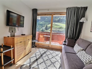 A room with a sofa, a TV, a wooden cabinet, a small table, and a view of a green landscape through a large window.