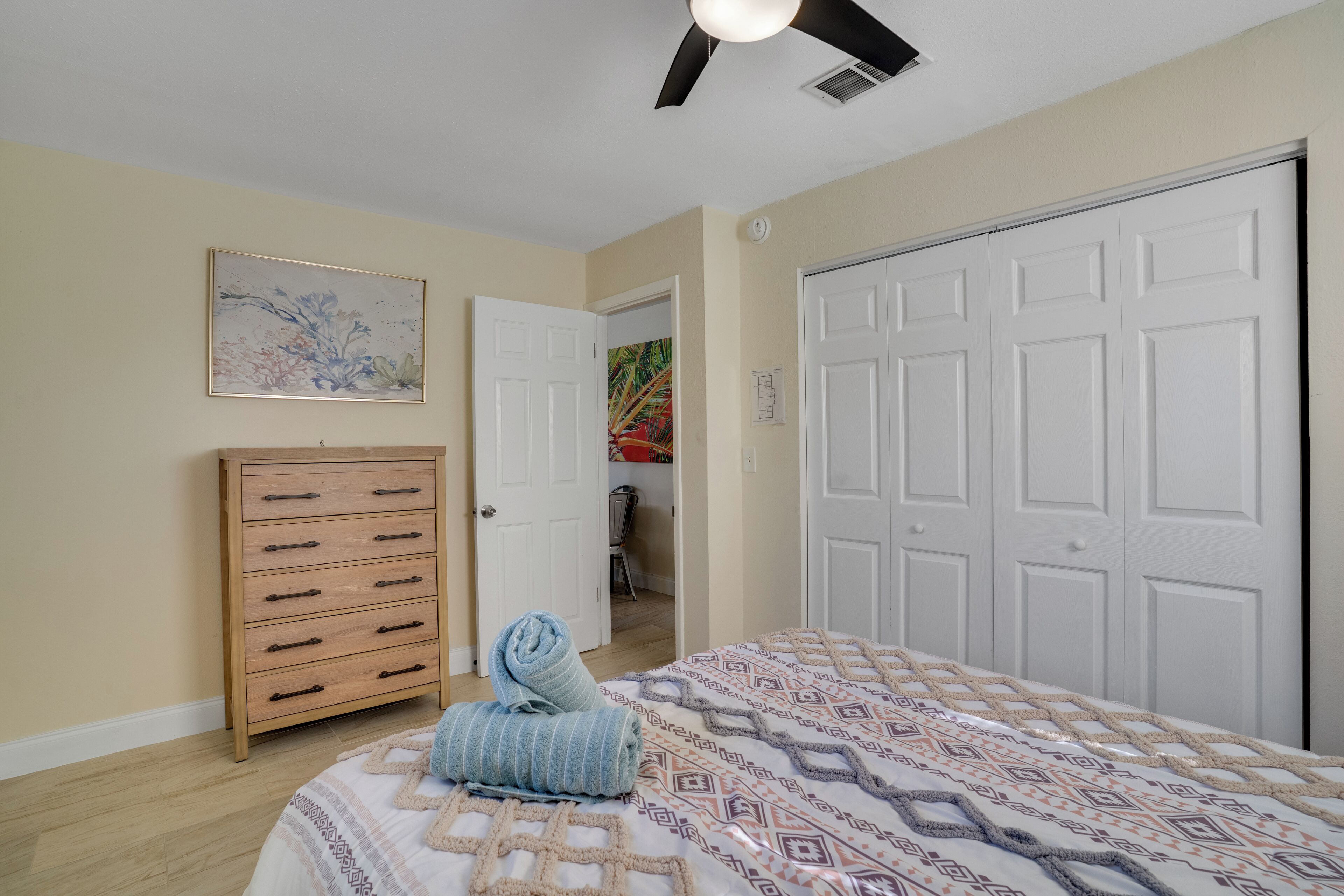 Une chambre à coucher comprenant un lit, une commode, un ventilateur de plafond et une porte donnant sur une autre pièce.