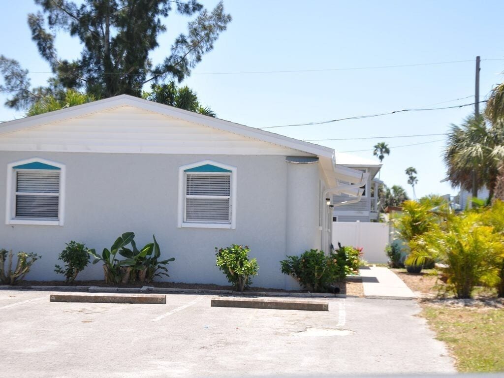 A single-story building with a flat roof, two windows with shutters, and a small front yard with various plants.
