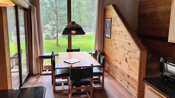 A dining area with a wooden table and chairs, large windows with curtains, and a view of a grassy yard. - Charming Ranch Cabin-New Premier Pool, 20 Minutes to Mt. Bachelor Lg Common Area (Sunriver)