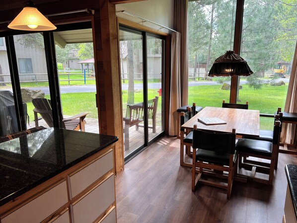 A modern kitchen with a large island, a dining area with wooden chairs and a table, and a sliding glass door leading to a backyard with a lawn and a wooden deck. - Charming Ranch Cabin-New Premier Pool, 20 Minutes to Mt. Bachelor Lg Common Area (Sunriver)
