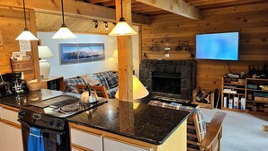 Ein gemütliches Blockhaus-Interieur mit Küchenbereich, der einen Herd, einen Wasserkocher und einen Esstisch umfasst. Ein Kamin mit Steinverkleidung und ein angebrachter Fernseher sind ebenfalls vorhanden.