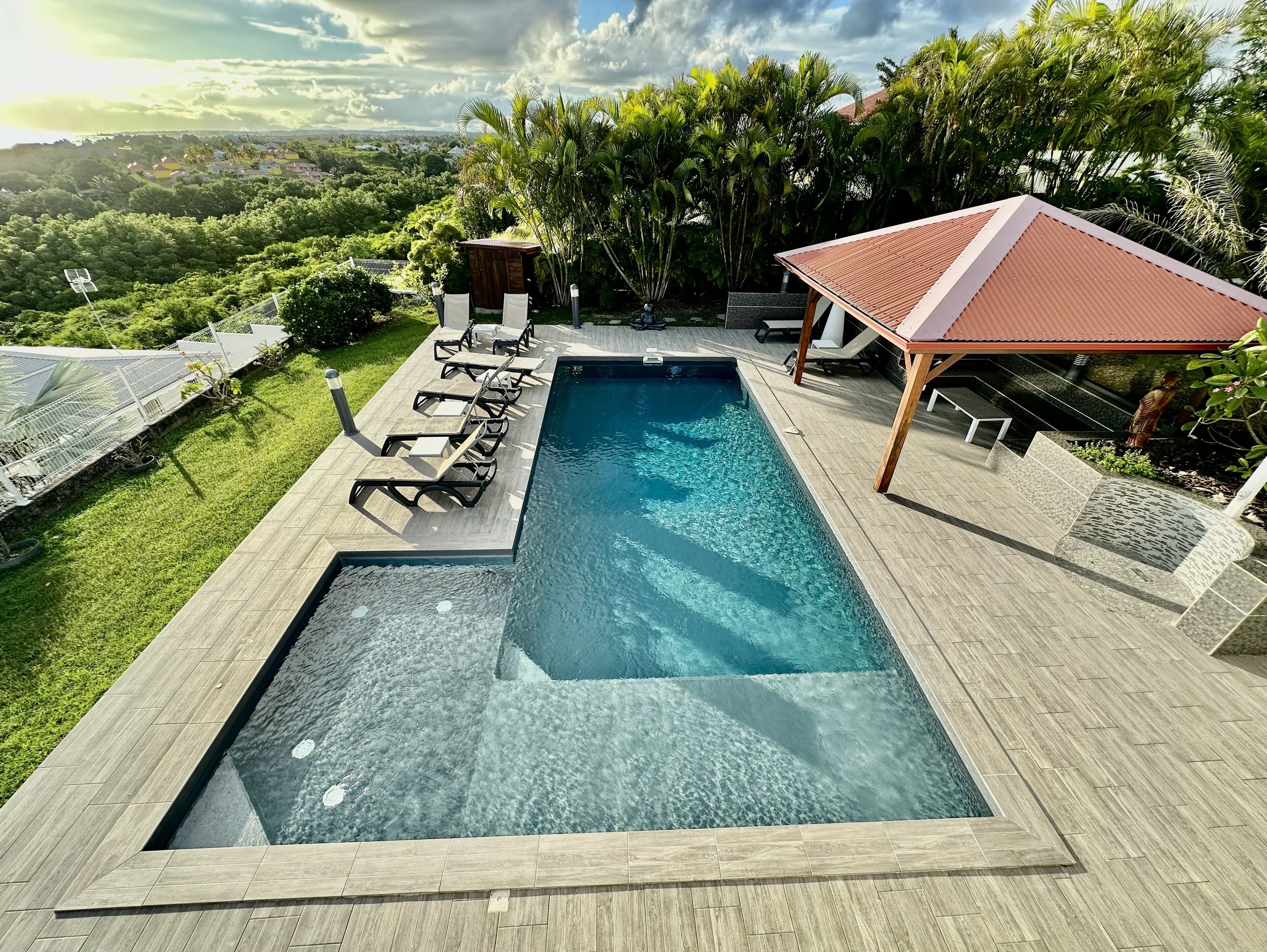 A rectangular swimming pool with a wooden deck, surrounded by lounge chairs and a covered seating area.