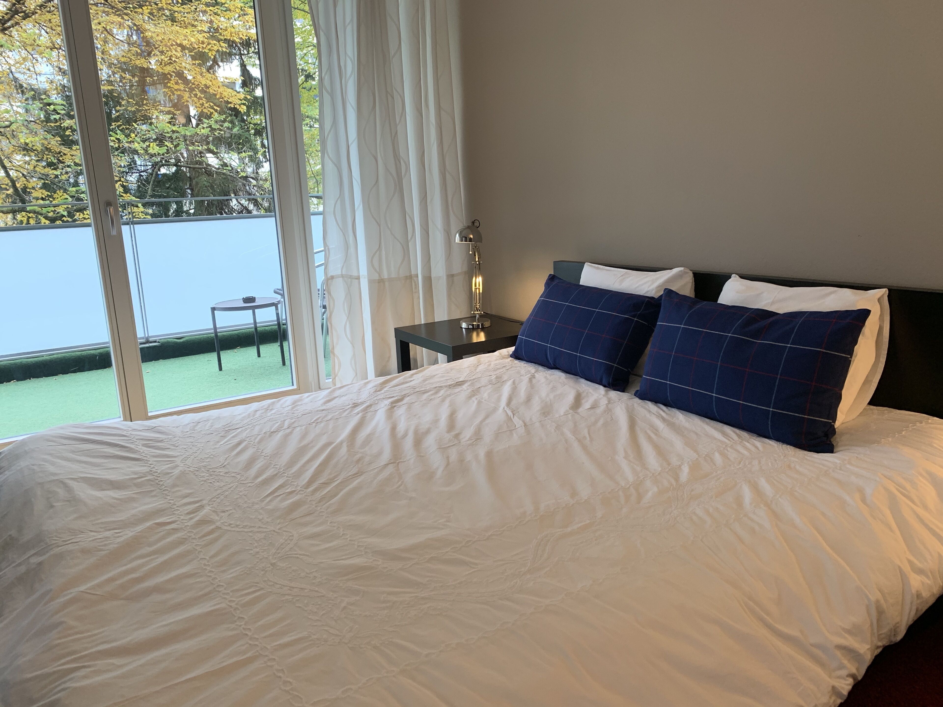 Ein Schlafzimmer mit einem großen Bett, einem Nachttisch, einer Lampe und einem Blick auf einen Balkon mit Tisch und Stühlen.
