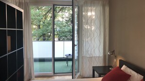 A hotel room with a bed, a nightstand, a lamp, a sliding glass door leading to a balcony with a table and chairs, and a view of trees. - Large charming apartment, bright, close to the center and the Old Town (Genève)