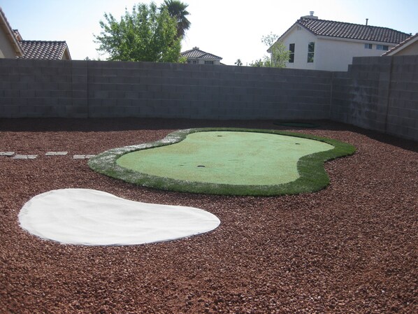 Un jardin avec une petite pelouse artificielle et une zone recouverte de gravier blanc, le tout entouré d’un mur de briques et de petits cailloux.