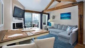 A modern living room with a grey sofa, a wooden dining table with placemats, a flat-screen TV, and a large window with a view of the sea. - Cozy Boat House Lake Charlevoix: Amazing View/Pool (Charlevoix)