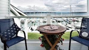 A balcony with a view of a marina, featuring a table with a striped tablecloth and two chairs with anchor-patterned cushions. - Cozy Boat House Lake Charlevoix: Amazing View/Pool (Charlevoix)