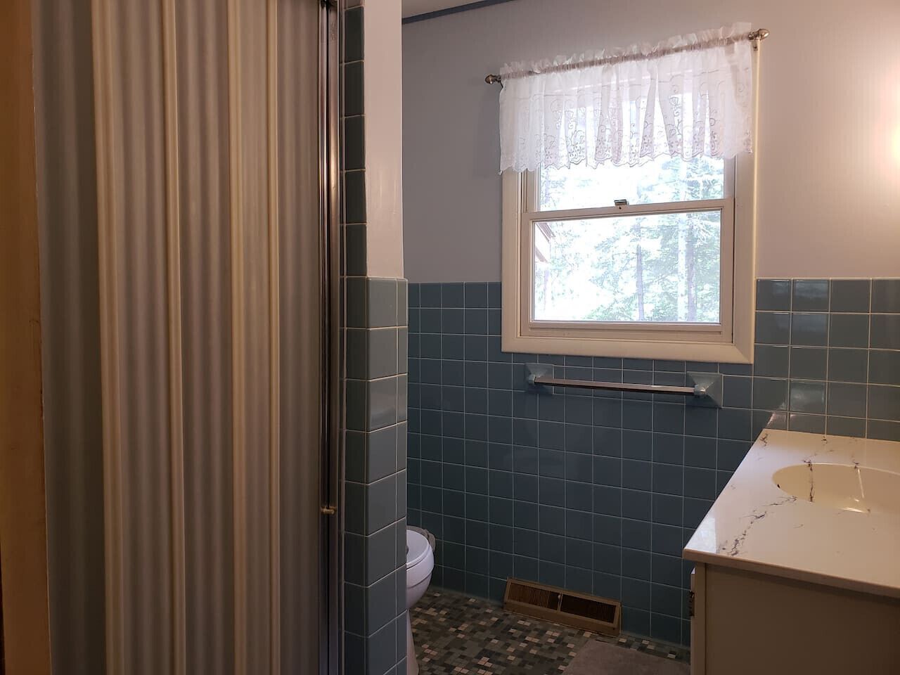 Une salle de bain avec des toilettes, une fenêtre ornée d’un rideau en dentelle et un lavabo avec un plan de travail en marbre.