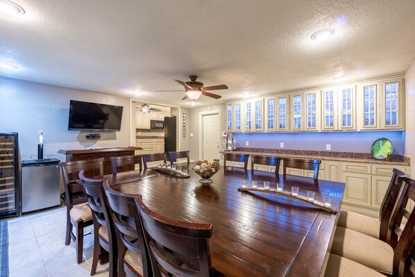 A spacious dining area with a wooden table and chairs, a ceiling fan, and a kitchen with light-colored cabinets and a large window.