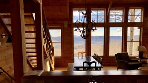 A wooden cabin interior with a dining area, a chandelier, and a view of the mountains through large windows. - Cabin With A View (Burnsville)