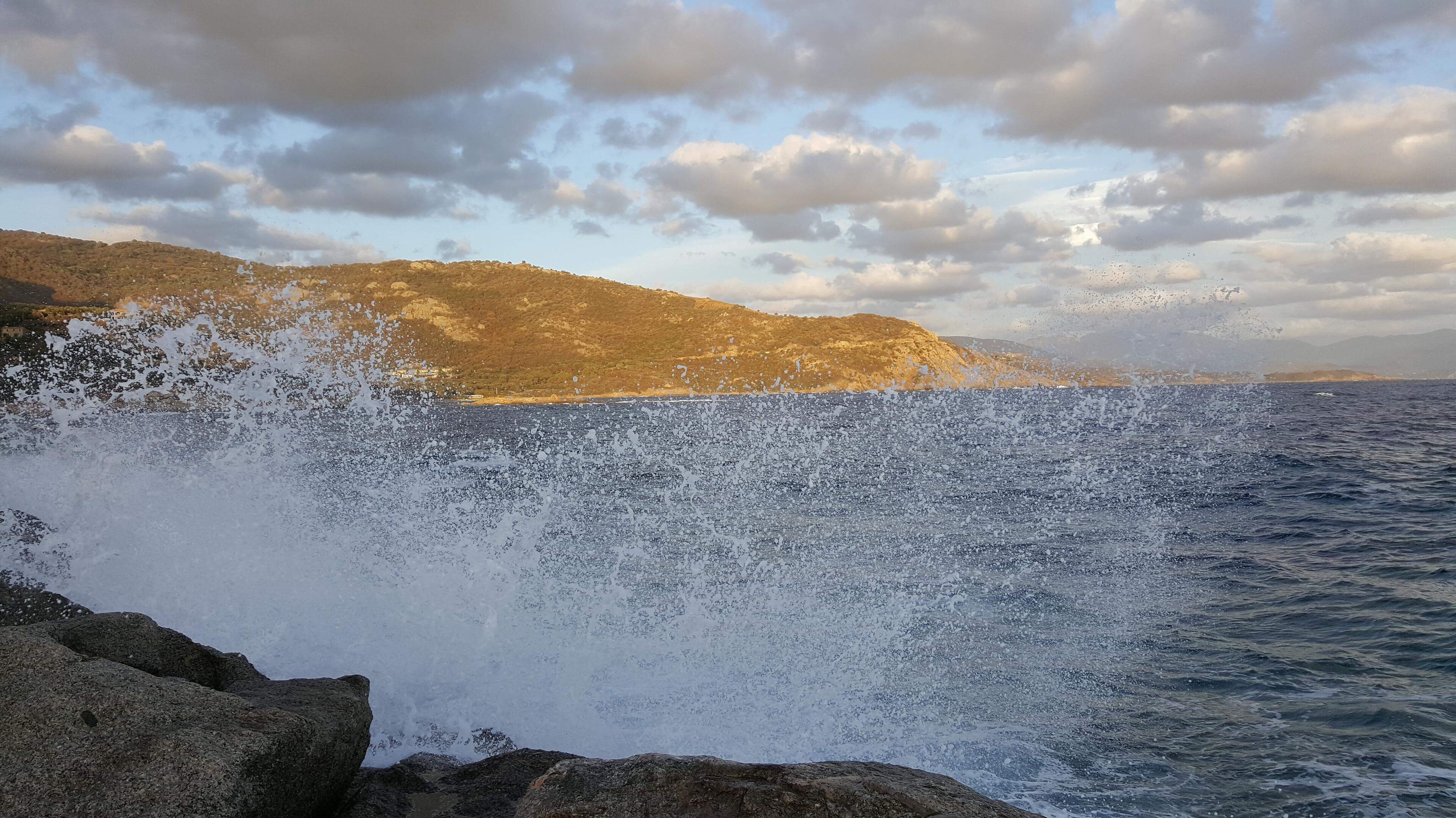 Escena costera con olas rompiendo contra rocas, un telón de fondo montañoso y cielo parcialmente nublado.