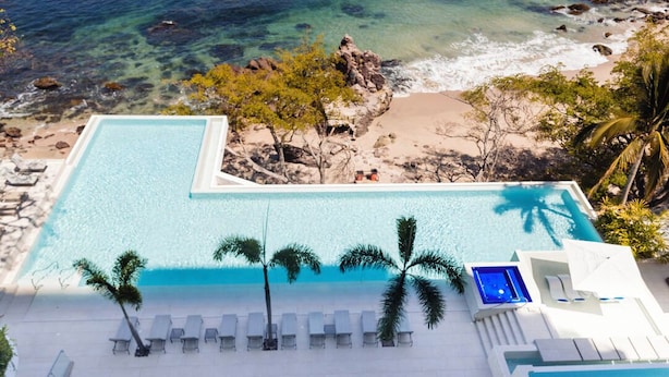 Una piscina infinita en la azotea con agua cristalina, rodeada de sillones y sombrillas, con vista a una playa con rocas y palmeras.