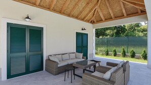 An outdoor patio with wicker furniture, a glass table, and a view of a green fence and trees. - Luxurious home in Hrvace (Hrvace)