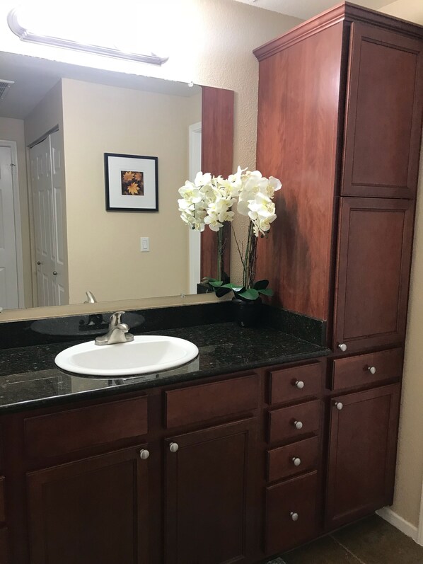 A bathroom with a dark wood vanity, a white sink, and a mirror above it. There is a framed picture on the wall and a vase with white flowers on the vanity.