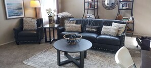 Un salon avec un canapé en cuir noir, une table ronde, une petite table d’appoint et une chaise. On y trouve également une bibliothèque et un miroir accroché au mur.