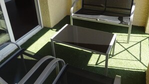 A balcony with a glass table and a cushioned bench, surrounded by artificial grass and a textured wall.