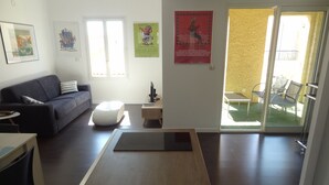 A modern living room with a sofa, a coffee table, and a TV. There are posters on the wall and a sliding glass door leading to an outdoor area with chairs.