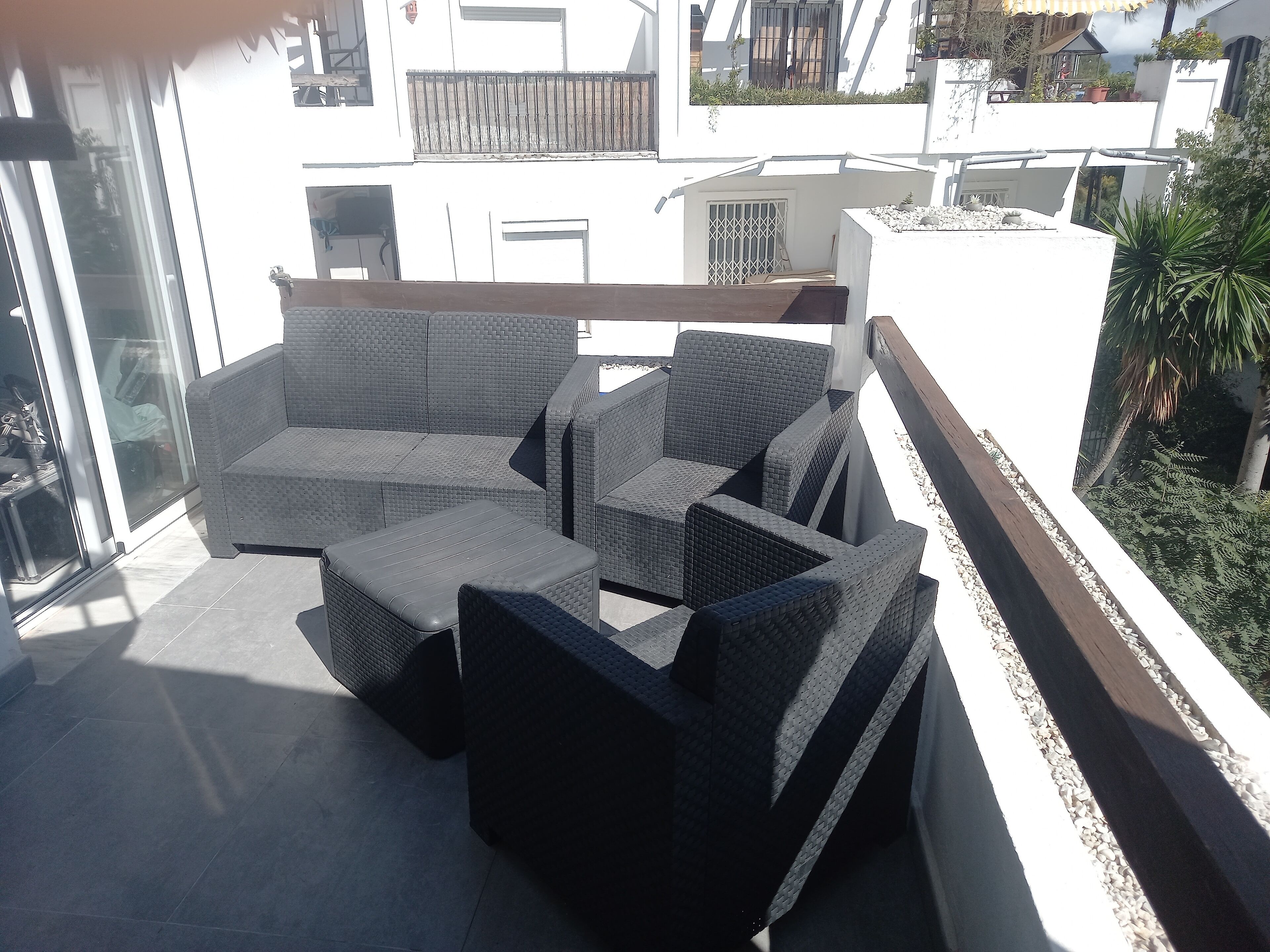 Un balcon avec un canapé et un pouf gris en fibres tressées, des murs blancs et une vue sur les bâtiments voisins.