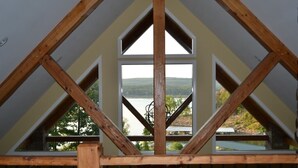 A room with a sloped ceiling, wooden beams, and a large window offering a view of a lake and hills. - Bellawood Resort #1 Lakefront Cabin with Boat Slip - Unobstructed View! (Greers Ferry)