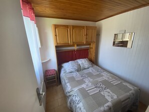A bedroom with a wooden wardrobe, a bed with a patterned bedspread, a small stool, and a mirror on the wall. - APARTMENT FOR 4 PEOPLE LOWER CHALET GARDEN FENCE BARBECUE PLACE QUIET (Saint-Pierre-Dels-Forcats)