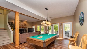 Un vaste salon doté d’une table de billard, de parquet, d’un escalier, d’une cheminée et d’une grande fenêtre donnant sur l’extérieur.