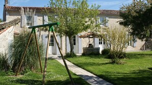 Un bâtiment traditionnel en pierre, avec un toit en tuiles, un jardin équipé d’une balançoire et une petite fontaine.