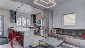 A modern kitchen with a central island, stainless steel appliances, and a grey sofa in the living area. - Woodside Ave - Close to Sundance Locations - Private Hot Tub (Park City)