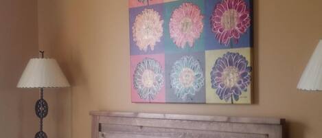 A bedroom with a bed, a headboard, a wall with a floral artwork, a lamp, and a nightstand.
