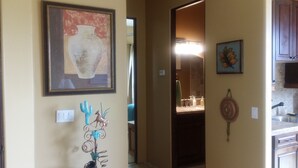 A hallway with a framed painting of a vase, a decorative metal piece, and a kitchen area with cabinets and a sink. - Vacation Rental Available (Scottsdale)