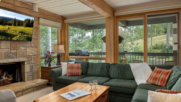 Ein gemütliches Wohnzimmer mit Kamin aus Stein, einem grünen Sofa, einem Holztisch und großen Fenstern mit Blick auf eine grüne Landschaft.