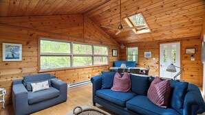 Ein gemütliches Blockhaus-Interieur mit Holzwänden, einem blauen Sofa, einem grauen Sessel und einem Essbereich mit Tisch und Stühlen.