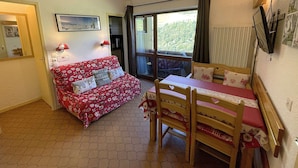 A cozy room with a red patterned sofa, a wooden dining table with chairs, a large window with a view of greenery, and a ceiling fan. - Cozy 4-Person Apartment, Ski-In/Ski-Out, West-Facing Balcony, Les Menuires (Les Menuires)