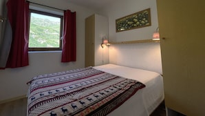 A bedroom with a bed, a window with red curtains, a wall-mounted lamp, and a painting. - Cozy 4-Person Apartment, Ski-In/Ski-Out, West-Facing Balcony, Les Menuires (Les Menuires)