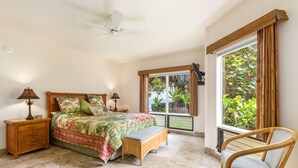 Une chambre à coucher comprenant un lit, des tables de chevet, un banc, une chaise, un ventilateur de plafond et une fenêtre donnant sur un paysage verdoyant.