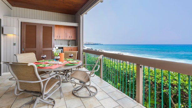 Un balcon avec une table dressée pour le dîner, offrant une vue sur une plage, l’océan et un paysage urbain lointain.