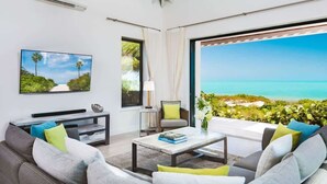 A living room with a TV, sofa, coffee table, and a view of the ocean. - Brise De Mer 6 BR Villa (Long Bay)