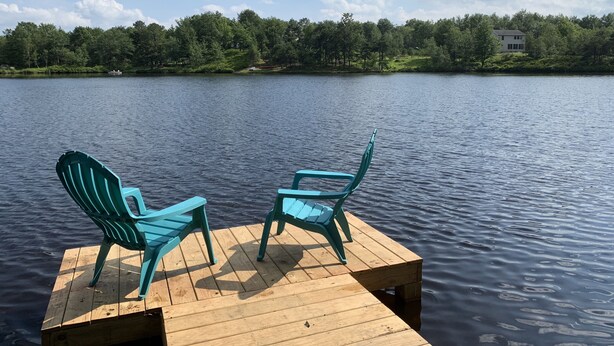 A wooden dock with two turquoise Adirondack chairs facing a calm lake, surrounded by lush greenery and a clear sky. - Beautiful Lakefront w Private Beach/Hot Tub/ Boats/ Fire Pit (Long Pond)