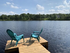 Un muelle de madera con dos sillas Adirondack turquesas mirando un lago tranquilo, rodeado de exuberante vegetación y un cielo despejado.