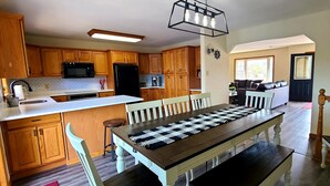 A modern kitchen with wooden cabinets, a central island, and a dining area with a checkered table runner. - Beautiful Lakefront w Private Beach/Hot Tub/ Boats/ Fire Pit (Long Pond)