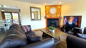 A living room with a sofa set, a coffee table, a fireplace, a television, and a dining area visible through an open door. - Beautiful Lakefront w Private Beach/Hot Tub/ Boats/ Fire Pit (Long Pond)