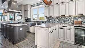 A modern kitchen with a large island, white cabinetry, a backsplash with blue and green tiles, and a hanging light fixture. - Fairwinds: Inviting Oceanfront Home Built to Entertain (Caswell Beach)