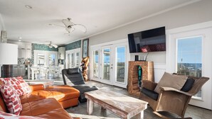 A spacious living area with a sofa, a coffee table, and a wicker chair. A television is mounted on the wall, and there is a view of the outdoors through large windows. - Fairwinds: Inviting Oceanfront Home Built to Entertain (Caswell Beach)