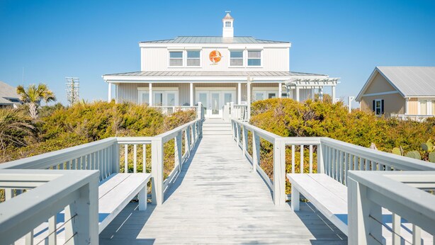 A white building with a central entrance, a covered porch, and a wooden walkway leading to it. - Fairwinds: Inviting Oceanfront Home Built to Entertain (Caswell Beach)