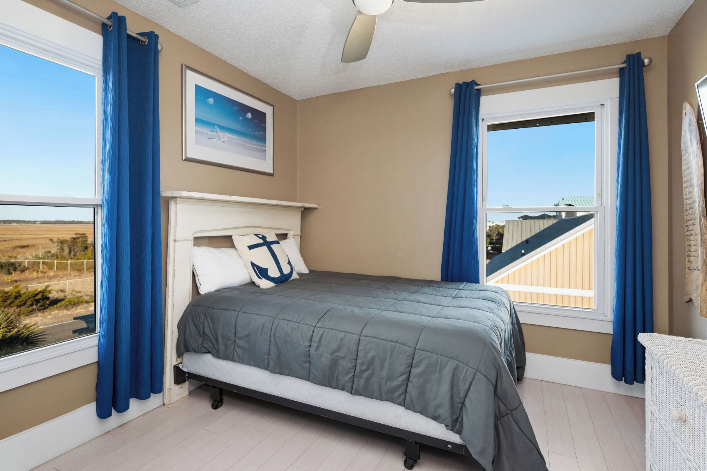 Une chambre à coucher avec un lit, un ventilateur de plafond, une fenêtre avec des rideaux, un tableau encadré et une vue sur l’extérieur.