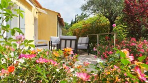 A garden with a yellow building, two white chairs, and blooming flowers.